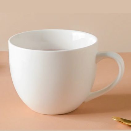 White ceramic mug on a peach surface with a small gold spoon nearby, empty inside.