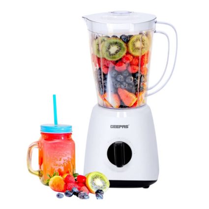 White blender with a pitcher filled with sliced kiwi, strawberries, blueberries, and other fruit; a smoothie jar with a blue lid and straw sits beside it.