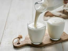 Two glasses of milk on a wooden paddle with milk being poured into the left glass from a pitcher, on a light wooden surface.