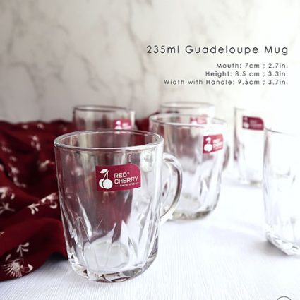 Clear glass mug with a red 'Red Cherry' logo on the front, labeled 235ml Guadeloupe Mug, on a white table with other mugs in the background.