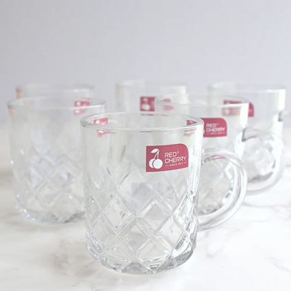 Clear glass mugs with a diamond-cut pattern lined up on a white marble surface, each bearing a red Red Cherry logo.”