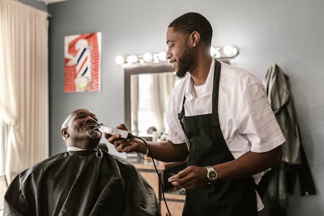 Barber in a white shirt and black apron trims a customer's beard with clippers as the client sits in a barber cape.