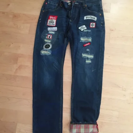 Pair of dark blue jeans with multiple fabric patches and distressed areas, laid flat on a wooden floor; cuffs rolled to show plaid lining.