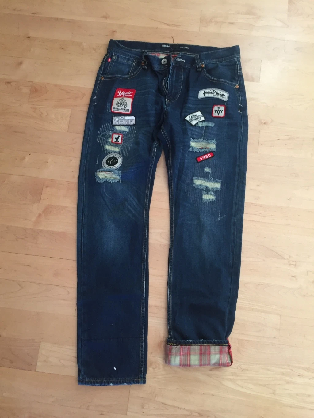 yesir jeans - Imavug Pair of dark blue jeans with multiple fabric patches and distressed areas, laid flat on a wooden floor; cuffs rolled to show plaid lining.